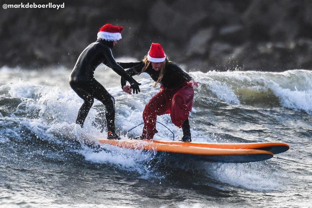 Two surfers wearing Santa hats ride a wave together on an orange surfboard.