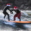 Two surfers wearing Santa hats ride a wave together on an orange surfboard.