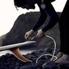 Surfer securing a leash to a surfboard on a rocky shoreline at dusk.