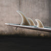Close-up of a surfboard with three fins against a concrete wall background.