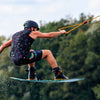 Man wakeboarding mid-air, wearing vibrant boardshorts and helmet, against a lush green backdrop.
