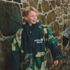 Child wearing White Water Kids Hard Shell Robe in camo near a stone wall.