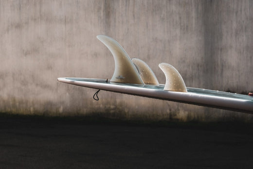 Close-up of a surfboard with three fins against a concrete wall background.