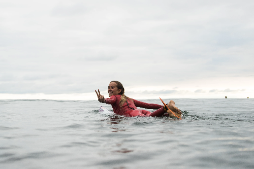 Surfer in a red wetsuit floating on calm ocean water, making a peace sign.