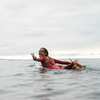 Surfer in a red wetsuit floating on calm ocean water, making a peace sign.