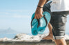 Man holding stylish blue flip-flops by the beach, showcasing casual men's footwear.