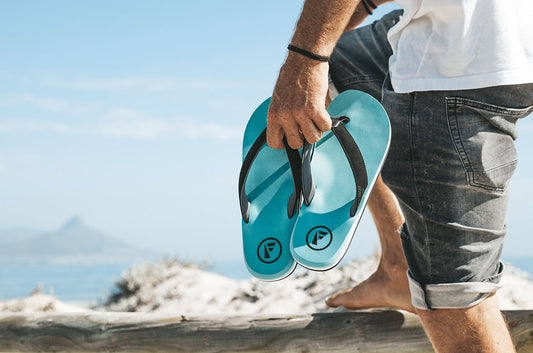 Man holding stylish blue flip-flops by the beach, showcasing casual men's footwear.