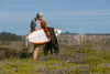 Surfer carrying a board and wetsuit through a grassy coastal landscape.