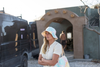 A woman wearing a pastel tie-dye bucket hat and matching tote bag stands outdoors near a van and an adobe-style building.