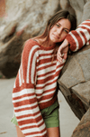 Cozy Sisstr Ocean Shores Sweater in rust, paired with green shorts near rocks.