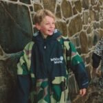 Child wearing White Water Kids Hard Shell Robe in camo near a stone wall.